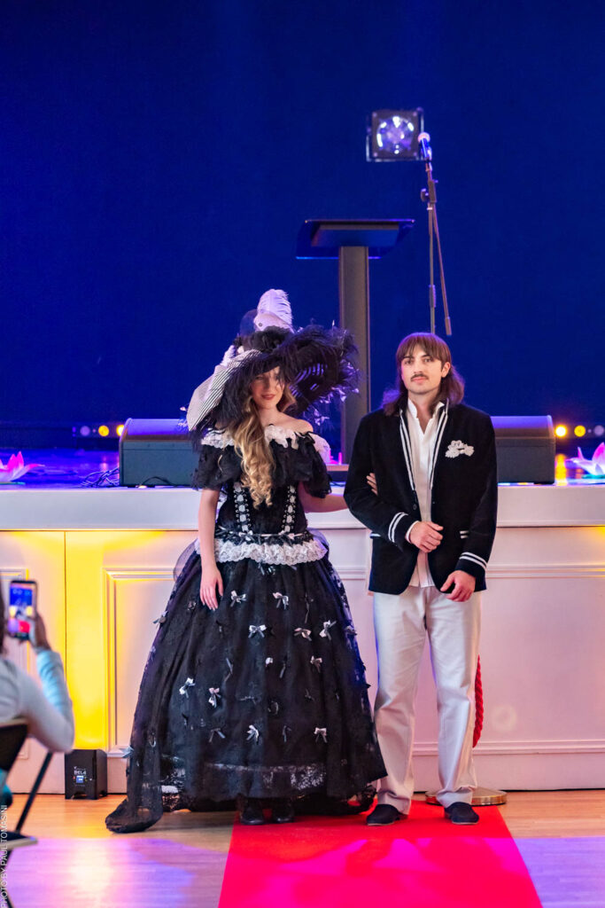 Ava, en corset lacé et jupon Belle Époque orné de nœuds, coiffée d'un chapeau à plumes, avance au bras de Paul, dandy en velours marine rayé.