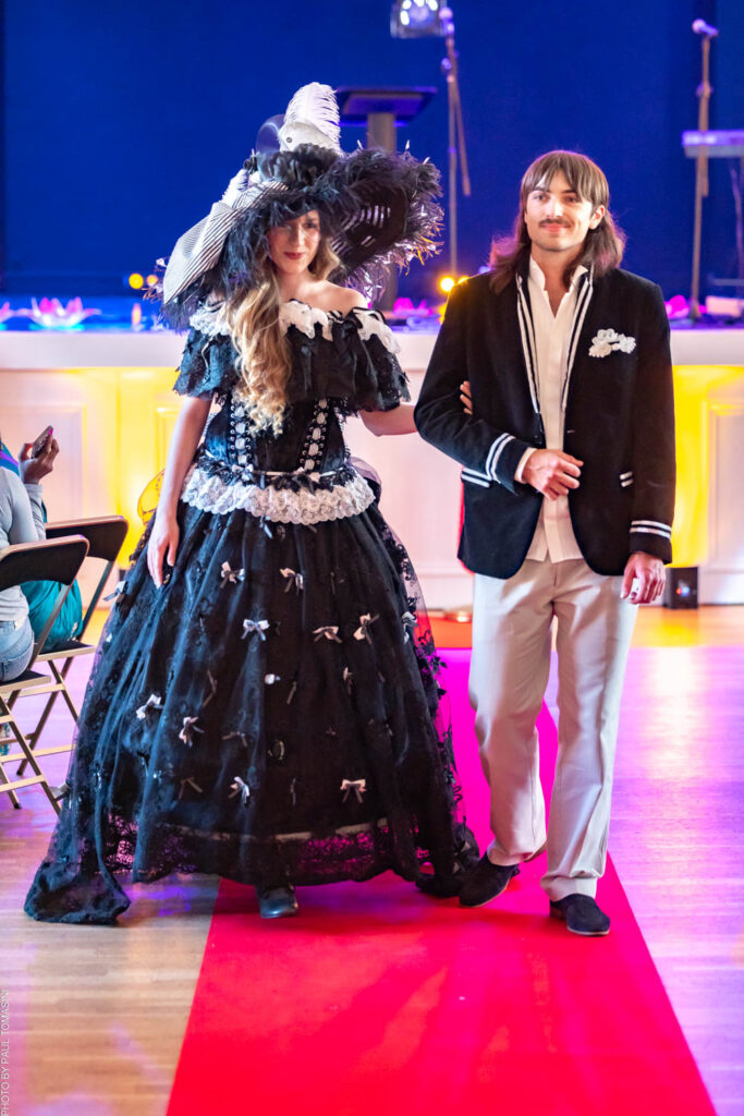 Chez Nathalie Froufrou, la mode se réinvente à partir de matières upcyclées : Ava sublime cette démarche dans un corset lacé et jupon Belle Époque orné de nœuds, coiffée d'un chapeau à plumes, au bras de Paul, dandy en velours marine rayé.