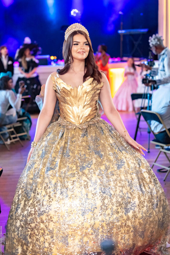 Couronnée d'un diadème étincelant, Ludivine défile dans une robe de bal dorée au corsage sculpté de plumes métalliques et à la jupe scintillante de paillettes argentées, une apparition royale digne d'un conte de fées haute couture.